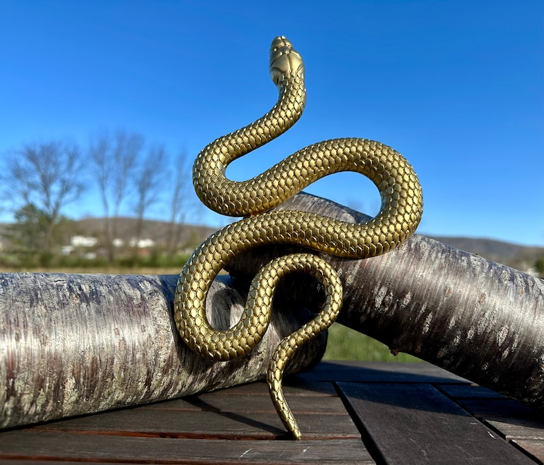 May include: A gold-colored snake sculpture with a textured surface, coiled around a piece of driftwood. The sculpture is on a wooden surface.