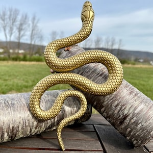 May include: A gold-colored snake sculpture with a textured, scaly surface. The snake is coiled in a relaxed pose, with its head raised and its tail curled underneath. The sculpture is resting on two pieces of tree bark.