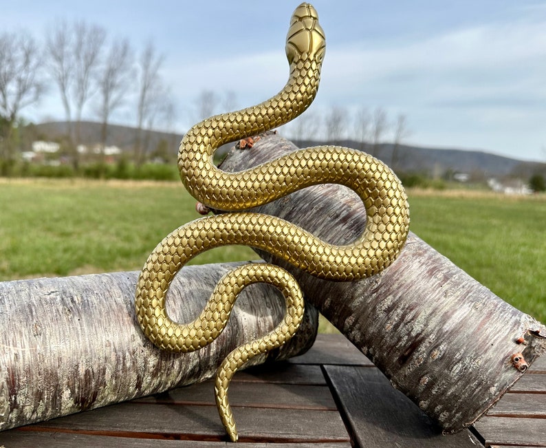 May include: A gold-colored snake sculpture with a textured, scaly surface. The snake is coiled in a relaxed pose, with its head raised and its tail curled underneath. The sculpture is resting on two pieces of tree bark.