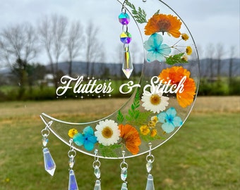 May include: A crescent moon-shaped resin ornament with pressed flowers and iridescent crystal accents. The ornament features a variety of flowers, including white daisies, orange marigolds, and blue forget-me-nots. The text "Flutters & Stitch" is etched into the resin.