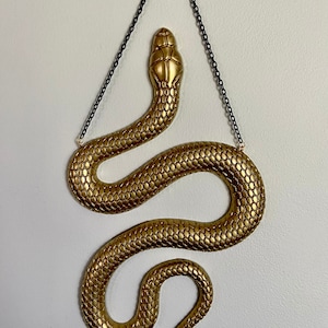 May include: A gold-colored snake wall hanging with a textured, scaly design. The snake is hanging from a black chain.