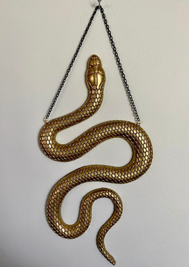 May include: A gold-colored snake wall hanging with a textured, scaly design. The snake is hanging from a black chain.