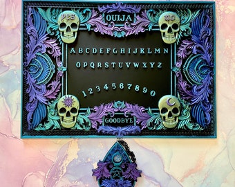 May include: A teal and purple framed Ouija board with a black planchette hanging below. The board features a skull design at each corner and the words "YES", "OUIJA", "NO", and "GOODBYE" along the top and bottom. The alphabet, numbers 1-9 and 0 are printed in white on the black board.