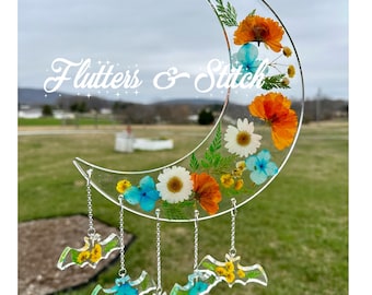 May include: A crescent moon-shaped resin ornament with pressed flowers and leaves in shades of orange, yellow, white, and blue.  The ornament has a silver chain with four bat-shaped charms hanging from it.  The text "Flutters & Stitch" is visible in the background.