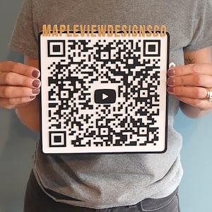 May include: A square black and white QR code with the text "MAPLEVIEWDESIGNSCO" in gold letters. The QR code has a YouTube play button in the center. The item is held by a person wearing a gray t-shirt.