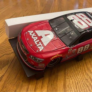 May include: A red and black Axalta racing car model with the number 88 prominently displayed. The model sits on a black stand, with the word "Axalta" and the number "4" visible on the hood. The car is on a wooden surface.