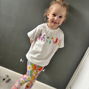 May include: A young child wearing a white t-shirt with the word "MINI" in colorful letters, paired with rainbow leggings featuring daisy patterns. The child is also wearing pink sneakers.