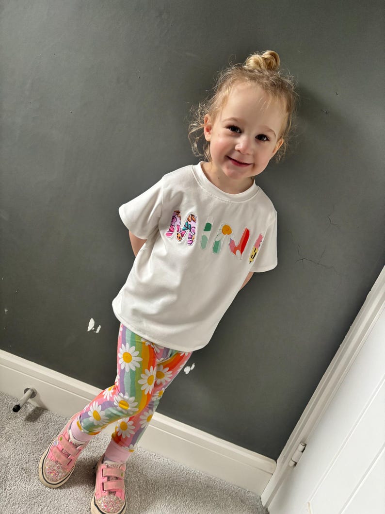 May include: A young child wearing a white t-shirt with the word "MINI" in colorful letters, paired with rainbow leggings featuring daisy patterns. The child is also wearing pink sneakers.