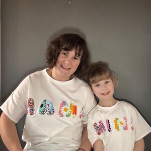 May include: Two people wearing matching white t-shirts with the word "MAMA" and "MINI" in colorful, floral-themed lettering. The shirts have a relaxed fit and short sleeves. The background is a solid gray.
