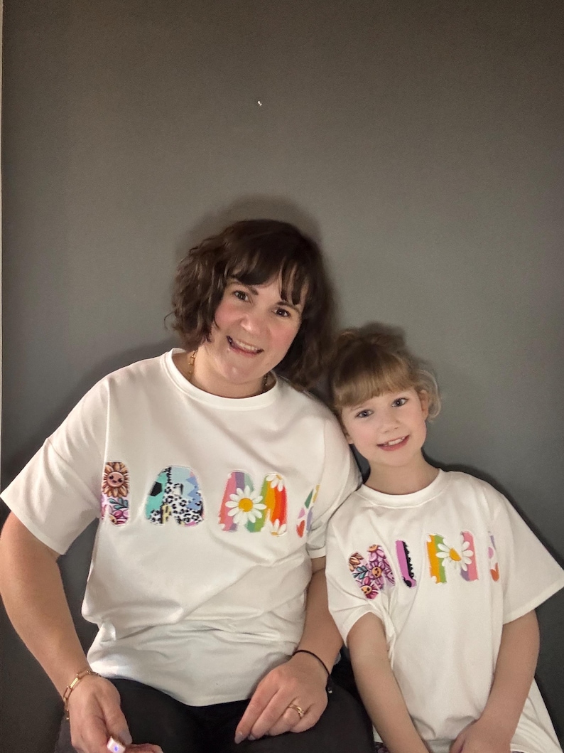 May include: Two people wearing matching white t-shirts with the word "MAMA" and "MINI" in colorful, floral-themed lettering. The shirts have a relaxed fit and short sleeves. The background is a solid gray.