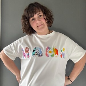 May include: White t-shirt with the word "MAMA" in colorful, patterned letters. The letters feature floral, animal print, and rainbow designs. The shirt has short sleeves and a relaxed fit.
