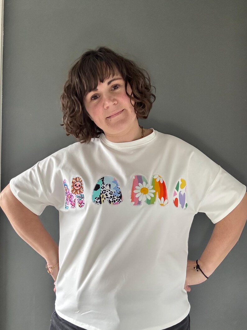 May include: White t-shirt with the word "MAMA" in colorful, patterned letters. The letters feature floral, animal print, and rainbow designs. The shirt has short sleeves and a relaxed fit.