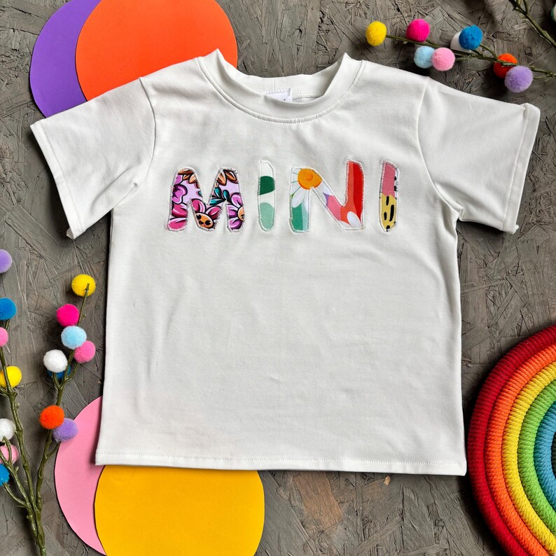 May include: White t-shirt with the word "MINI" in colorful, floral-patterned letters. The letters are in pink, green, red, and yellow. The shirt has short sleeves and a crew neck.