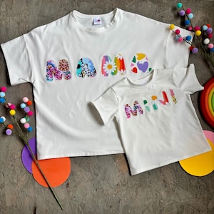 May include: Two white t-shirts, one adult and one child size, with the words "MAMA" and "MINI" appliqued on the front. The letters are decorated with colorful floral and patterned fabrics. The shirts are short-sleeved.