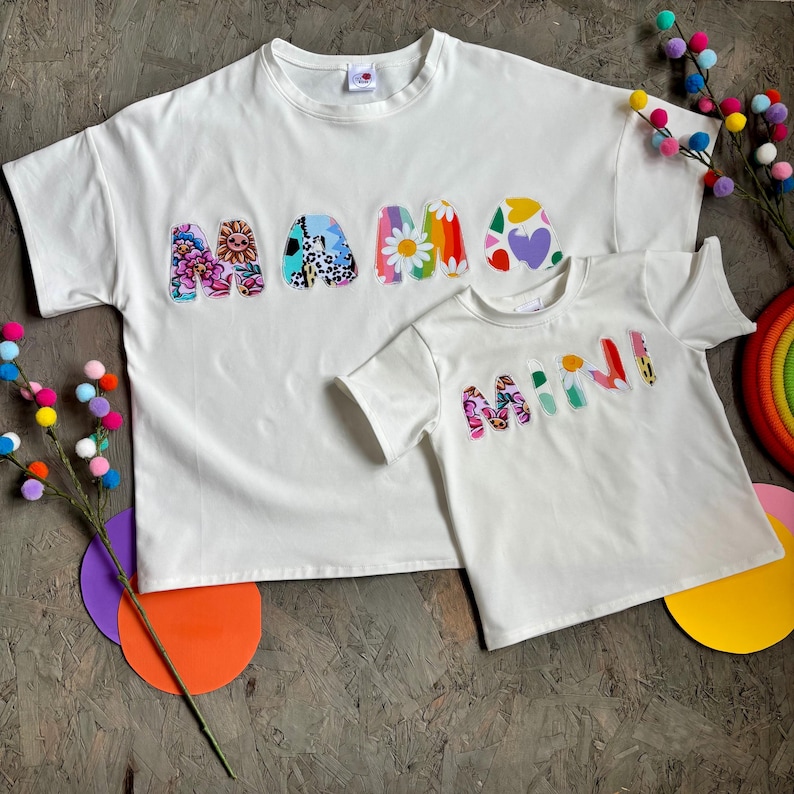 May include: Two white t-shirts, one adult and one child size, with the words "MAMA" and "MINI" appliqued on the front. The letters are decorated with colorful floral and patterned fabrics. The shirts are short-sleeved.
