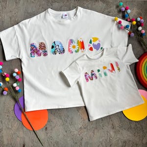 May include: Two white t-shirts, one adult and one child size, with the words "MAMA" and "MINI" appliqu&eacute;d in colorful floral and patterned fabric. The shirts are short-sleeved and have a casual fit. Decorative pom-poms and a rainbow are in the background.