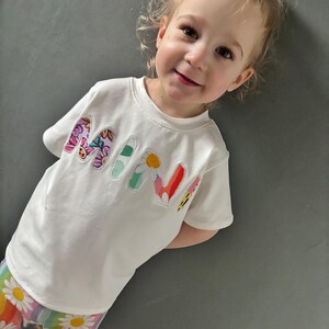 May include: A white t-shirt with the word "MINI" in colorful floral letters. The child is also wearing rainbow-striped pants with white daisy patterns.