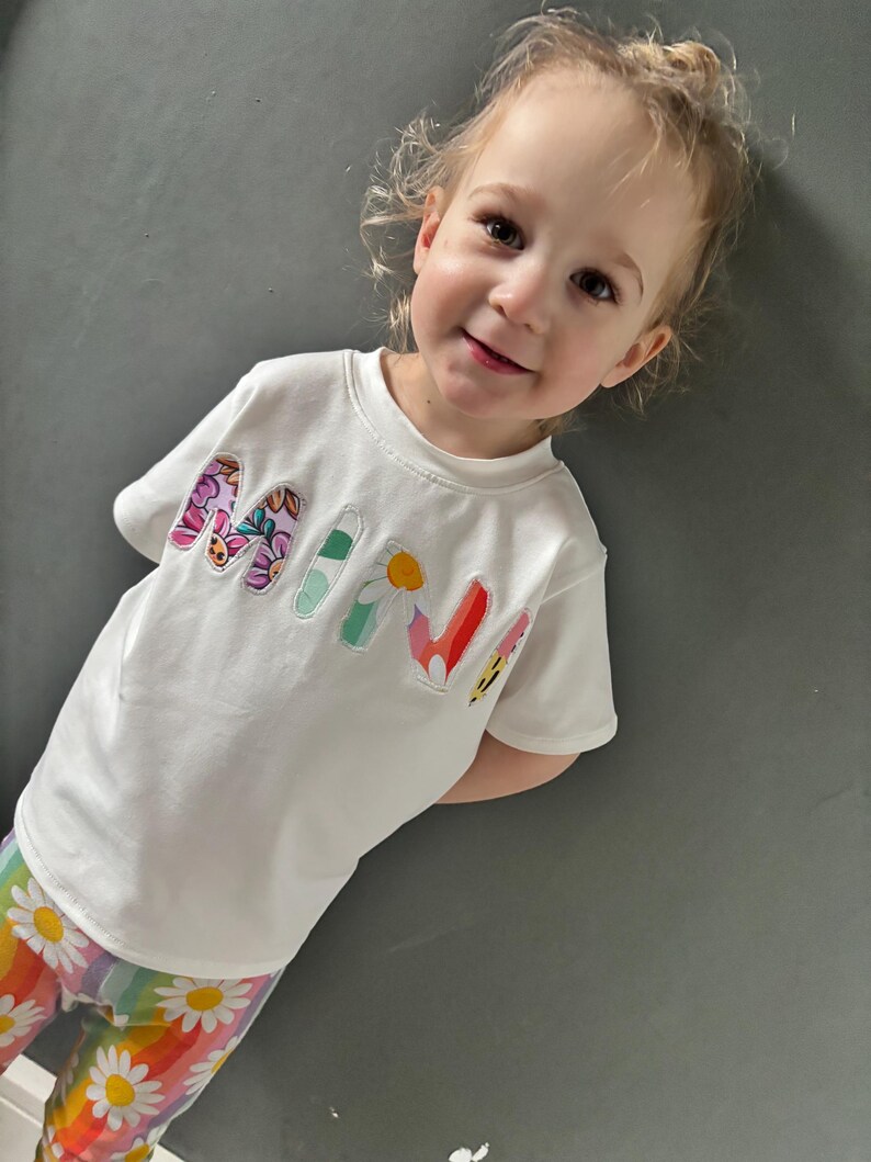 May include: A white t-shirt with the word "MINI" in colorful floral letters. The child is also wearing rainbow-striped pants with white daisy patterns.