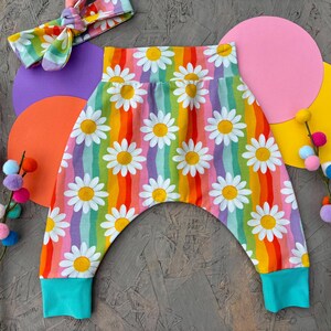 May include: Baby trousers with a rainbow stripe pattern and white daisy flowers, alongside a matching headband. The cuffs are turquoise. Brightly coloured paper circles and pom-pom stems are arranged around the clothing on a grey surface.