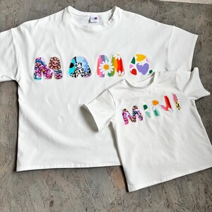 May include: Two white t-shirts, one adult and one child size, with the words "MAMA" and "MINI" appliqued on the front. The letters are decorated with colorful floral and abstract patterns. The shirts are made of a soft material.