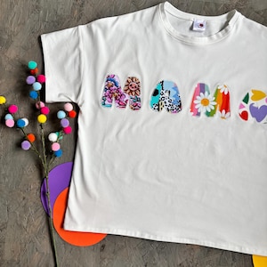 May include: White t-shirt with the word "MAMA" in colorful, patterned letters. The letters feature floral, animal print, and heart designs. A decorative branch with colorful pom-poms and paper cutouts are in the background.