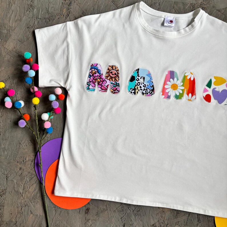 May include: White t-shirt with the word "MAMA" in colorful, patterned letters. The letters feature floral, animal print, and heart designs. A decorative branch with colorful pom-poms and paper cutouts are in the background.