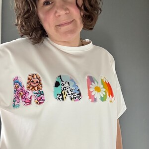 May include: White t-shirt with the word "MAM" in colorful, patchwork-style letters. The letters feature floral, sunflower, and rainbow daisy designs. The shirt has a crew neck and short sleeves.