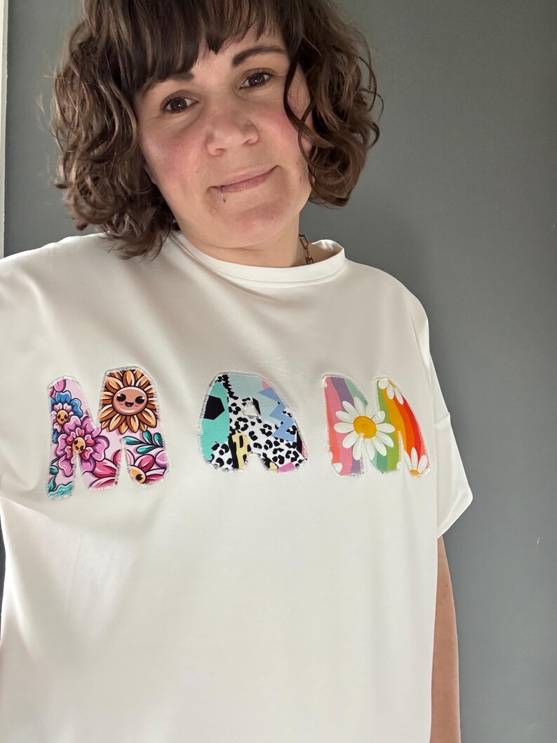 May include: White t-shirt with the word "MAM" in colorful, patchwork-style letters. The letters feature floral, sunflower, and rainbow daisy designs. The shirt has a crew neck and short sleeves.