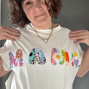 May include: White t-shirt with the word "MAMA" in colorful, floral, and patterned letters. The letters feature a sunflower, daisies, and other designs. The shirt is a casual, everyday wear item.