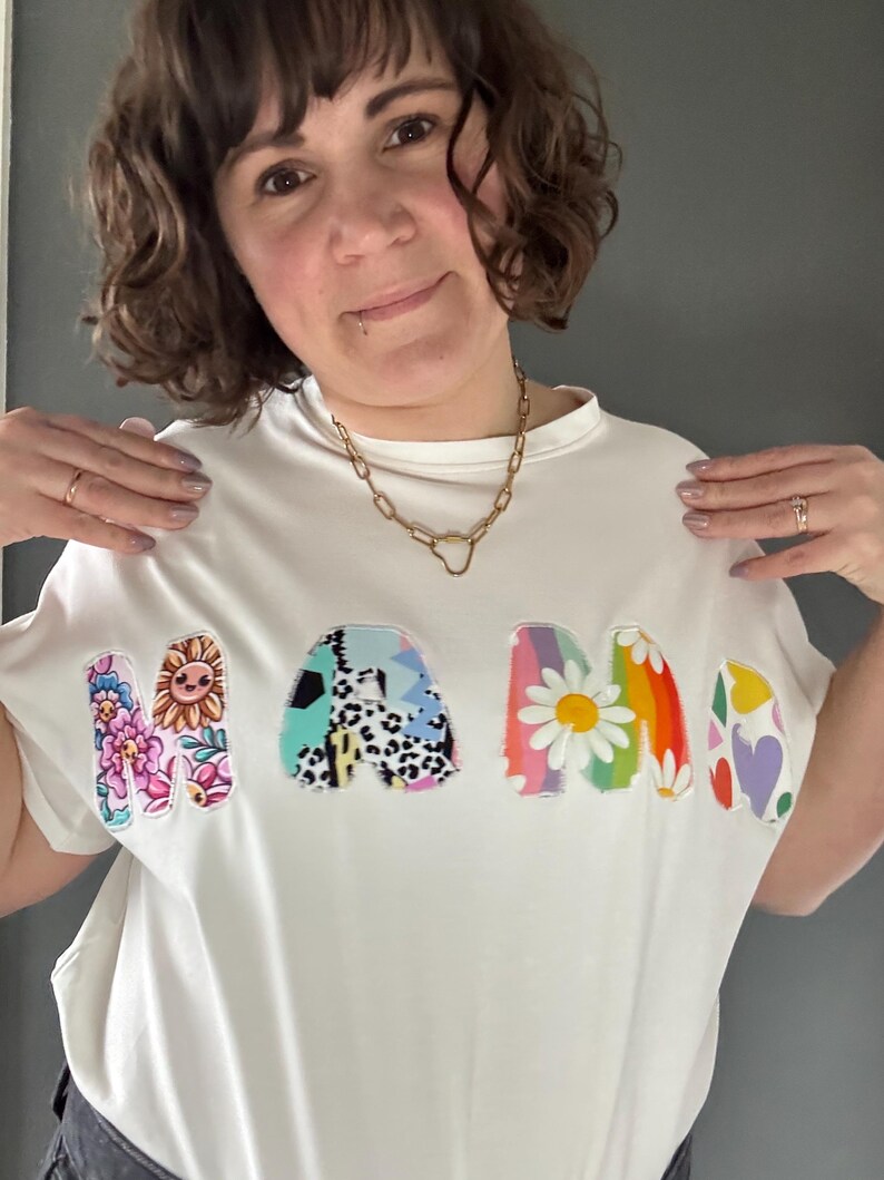 May include: White t-shirt with the word "MAMA" in colorful, floral, and patterned letters. The letters feature a sunflower, daisies, and other designs. The shirt is a casual, everyday wear item.