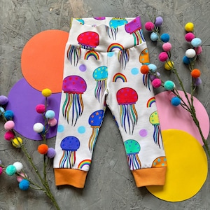 May include: Baby leggings in white, patterned with colourful jellyfish, rainbows, and stars. The leggings have an orange cuff and a high waistband. The jellyfish are in various colours. The background is light grey.