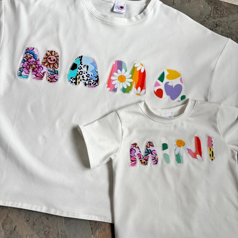 May include: Two white t-shirts, one adult and one child size, with the words "MAMA" and "MINI" appliqued on the front. The letters are in a colorful floral and patterned design. The shirts are made of a soft material.