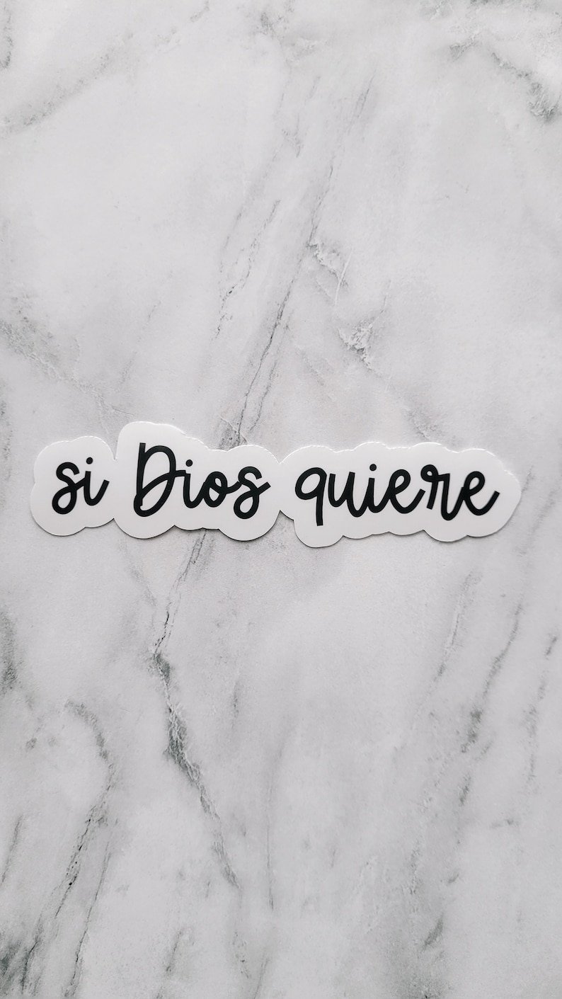 May include: White sticker with black lettering that reads "si Dios quiere" on a white marble background.
