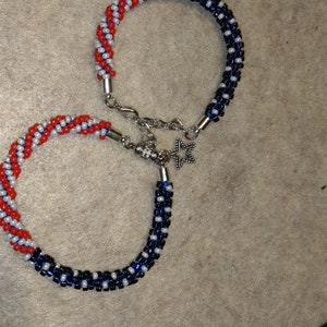 May include: Two beaded bracelets with red, white, and blue color patterns. The bracelets are connected with a silver clasp and a star charm.
