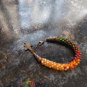 May include: A beaded bracelet with a gradient of green, orange, and red beads. The bracelet has a bronze clasp with a leaf charm.