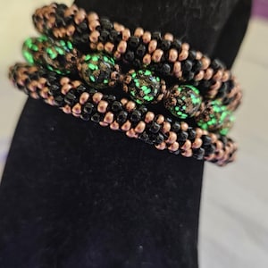 May include: Three beaded bracelets. The bracelets are black and copper-colored, with a central row of green and black beads. The bracelets are stacked on top of each other, creating a layered effect. The beads have a speckled appearance.