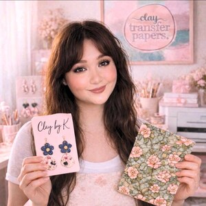 May include: A woman holding a card with "Clay by K" and flower earrings, and a floral patterned paper. The earrings feature blue and floral designs. The paper has a green background with pink flowers. A sign in the background reads "clay transfer papers."