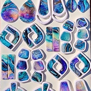May include: An assortment of handmade resin jewelry components in various shapes, including teardrops, rectangles, and geometric designs. The pieces feature swirling patterns in shades of blue, teal, purple, and white, with a glittery, iridescent finish. Each piece is outlined in white.