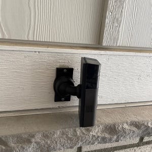 May include: A black rectangular security camera mounted on a wall with a circular base and an adjustable arm. The device features a smooth, dark finish with a vertical grille pattern on the side and a glossy lens area on the front facing outward.