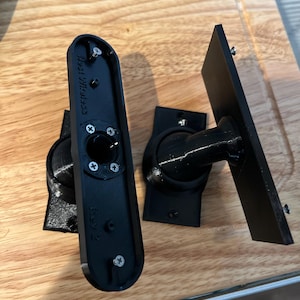 Puede incluir: Soportes de montaje de plástico negro con tornillos. Un soporte tiene el texto "Nest Wireless Rev 2". Los soportes están diseñados para sujetar un dispositivo. Los soportes están sobre una superficie de madera.