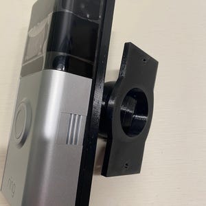 Puede incluir: Un timbre de video plateado y negro con un botón circular y la palabra ring impresa en la parte inferior. El dispositivo está unido a un soporte de montaje negro, rectangular e impreso en 3D con una abertura circular central y dos orificios.
