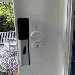 Może przedstawiać: Biały system dzwonka do drzwi z czarnym wierzchem i srebrnym korpusem jest zamontowany na białej ramie drzwi. Białe, okrągłe pokrętło z tarczą jest zamontowane obok dzwonka. W tle widać werandę z wzorzystym dywanem i białą balustradą.
