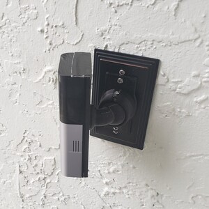 Puede incluir: Un timbre con cámara inteligente rectangular en color negro y plateado montado sobre una placa de pared rectangular negra. El dispositivo cuenta con una lente de cámara vertical, una rejilla de altavoz inferior y un brazo ajustable negro.