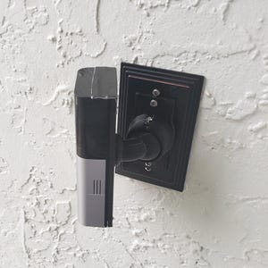 Puede incluir: Un timbre con cámara inteligente rectangular en color negro y plateado montado sobre una placa de pared rectangular negra. El dispositivo cuenta con una lente de cámara vertical, una rejilla de altavoz inferior y un brazo ajustable negro.