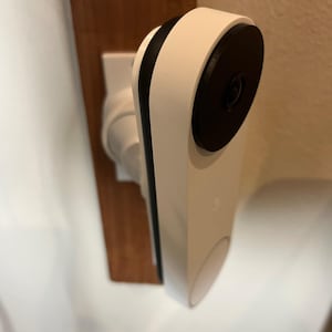 Op de afbeelding: Een witte slimme deurbel met een zwarte cameralens en zwarte rand. De deurbel is gemonteerd op een houten oppervlak en een witte muur. De deurbel heeft een afgeronde bodem en een luidspreker.