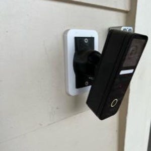 Puede incluir: Una cámara de seguridad rectangular negra montada en un soporte blanco y negro, fijada a una pared beige. La cámara tiene una pequeña franja blanca y una luz roja. El soporte es rectangular y tiene un brazo curvo.