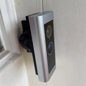 May include: A rectangular video doorbell with a silver metallic finish and a black front panel. The device features a circular camera lens at the top and a round button below it, mounted on a black bracket attached to a white door frame.