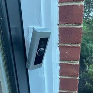 May include: A silver rectangular doorbell camera mounted on a white surface next to a red brick wall. The camera has a black face with a circular button and a lens.