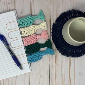 May include: Six colorful crocheted bookmarks with fringe tassels. The bookmarks are attached to gold metal clips. The bookmarks are arranged in a row on a white notebook.