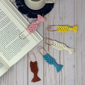 May include: Five colorful macrame fish bookmarks with gold metal clips. The bookmarks are pink, yellow, white, blue, and brown.